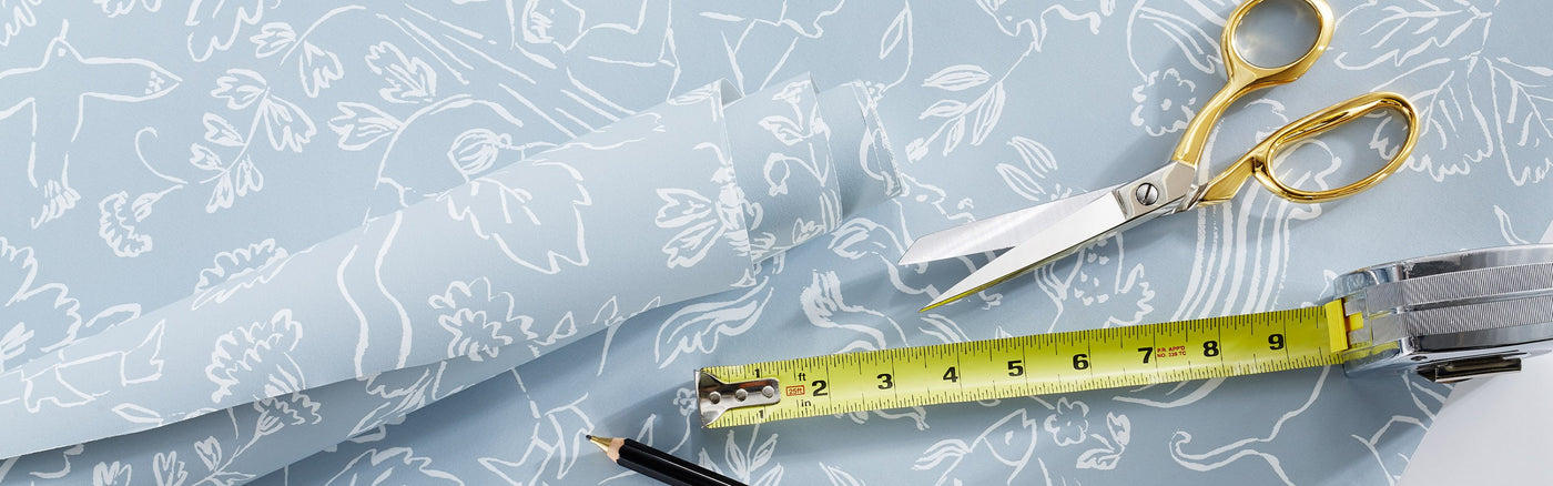 A light blue wallpaper with a white floral pattern with a roll of the same wallpaper laying on top, gold-handled scissors, a black pencil and a tape measure partially extended.